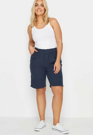 Lächelnde Frau mit blondem Haar, die ein weißes ärmelloses Oberteil, marineblaue Shorts mit elastischem Bund und weiße Sneakers trägt und mit einer Hand in der Tasche steht.