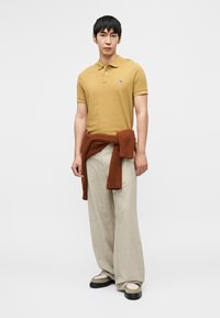 Polo jaune moutarde à manches courtes, associé à un pantalon beige clair, ample, et un pull marron côtelé noué autour de la taille. Chaussures noires.