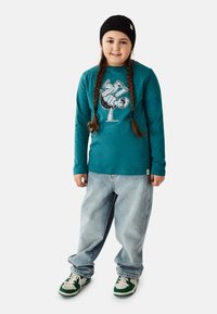 Langærmet skjorte i blågrøn med grafisk tryk, kombineret med lyseblå, løstsiddende jeans og sneakers med grønne detaljer, kompletteret med en sort strikket hue.