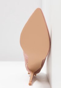 Chaussure à talon haut nude à bout pointu vue de la semelle, positionnée verticalement contre un mur blanc et un sol blanc.