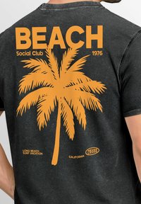 Black cotton t-shirt featuring an orange palm tree graphic, large text reading "BEACH Social Club 1976," and additional text detailing location and brand.
