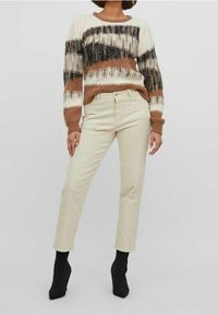 Gestreifter Strickpullover mit braunen, cremefarbenen und schwarzen Mustern, kombiniert mit hellbeigen Hosen und schwarzen Stiefeletten mit Absatz. Weiche Textur, lässige Passform.