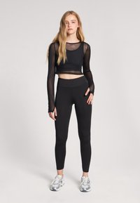 Czarna, krótka bluzka z długim rękawem i siateczkowymi detalami, połączona z czarnymi legginsami z wysokim stanem. Model nosi srebrne buty sportowe.