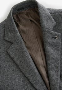 Blazer en laine gris chevrons avec doublure intérieure marron à motifs et une épingle de revers argentée en forme de couronne sur le côté gauche.