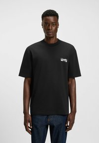 Sort bomulds T-shirt med korte ærmer og rund hals, med et hvidt logo på venstre bryst. Denim jeans bæres nedenunder.