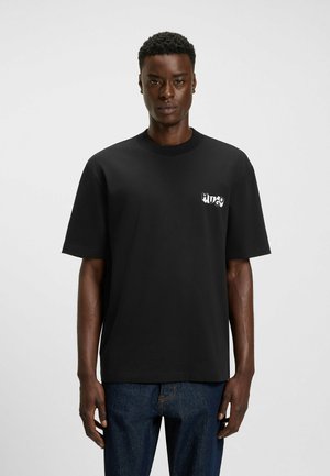 Schwarzes Baumwoll-T-Shirt mit kurzen Ärmeln und Rundhalskragen, mit einem weißen Logo auf der linken Brust. Darunter werden Jeans aus Denim getragen.