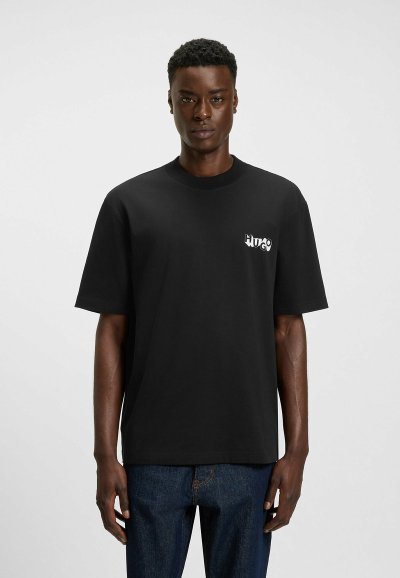 Sort bomulds T-shirt med korte ærmer og rund hals, med et hvidt logo på venstre bryst. Denim jeans bæres nedenunder.