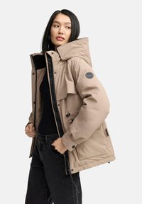Beige padded jacket with a high collar, large front pockets, and zipper closure, worn over a black top and paired with black jeans.