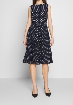 Mekko, joka on syvän sininen, hihaton, valkoisilla pilkuilla, vartalonmyötäinen yläosa, vyötäröltä kireä ja liehuva, laskostettu polveen ulottuva hame. Mustat, kärjet ylöspäin osoittavat korkokengät.