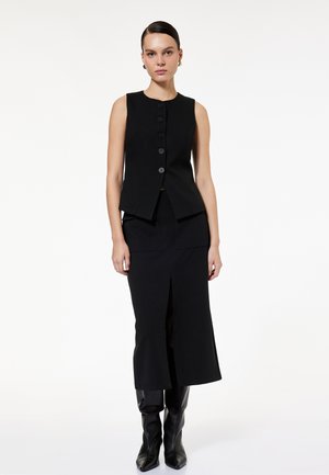 Kvinde iført sort ærmeløs knappet vest, sort midinederdel med frontslids og sorte knæhøje støvler, stående foran en hvid baggrund.