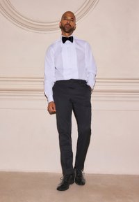 Camicia bianca con davanti a pieghe, papillon nero, pantaloni scuri e scarpe nere lucide. La figura è in piedi contro uno sfondo chiaro e strutturato.