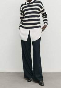 Personne portant un pull rayé noir et blanc par-dessus une chemise blanche, un pantalon large noir à fines rayures et des chaussures noires, debout les mains dans les poches.