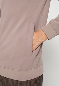 Taupe ritssluitingjack gemaakt van gestructureerde stof, voorzien van een zijzak en geribbelde manchetten, ontworpen voor een relaxte pasvorm.