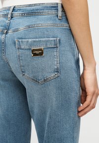 Ανοιχτό μπλε τζιν με καμπυλωτή πίσω τσέπη που διαθέτει μεταλλική ετικέτα μάρκας και αντίθετη ραφή· μαλακή υφή, ίσια εφαρμογή.