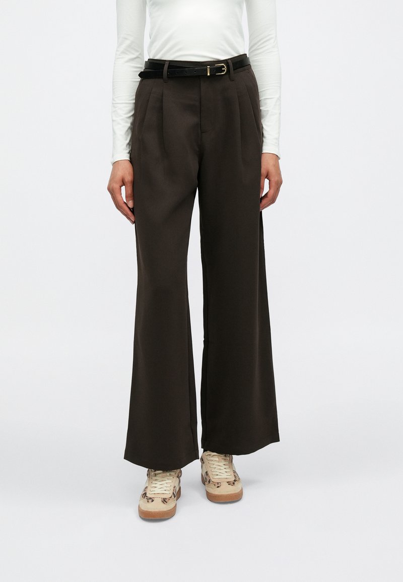 Brown, wide-leg trousers with pleats, made of smooth fabric. Black belt with gold buckle. Worn with beige, patterned sneakers.