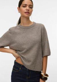 Femme aux cheveux bruns attachés en queue de cheval, portant un pull gris à manches courtes, un jean foncé, des boucles d'oreilles créoles et pendantes, ainsi que trois bracelets épais.