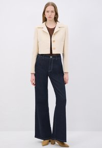 Beige kort tweedjacka med knappstängning och två fickor, tillsammans med mörk denim med utsvängda byxor som har tydlig sömdetaljering.