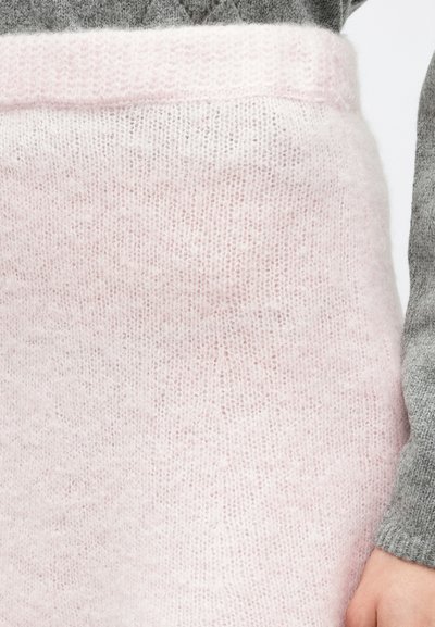 Svetloružový pletený sveter s mäkkou textúrou. Má široký rebrovaný výstrih. Nosený cez sivý textúrovaný materiál.