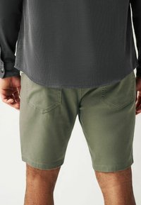 Shorts en coton verts avec une texture lisse et deux poches arrière, associés à une chemise à manches longues foncée avec un motif léger en côte.