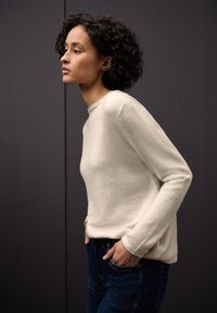 Jasnobeżowy sweter z długim rękawem o okrągłym dekolcie, wykonany z miękkiego materiału. Model nosi ciemnoniebieskie dżinsy, ręce w kieszeniach, profil boczny.