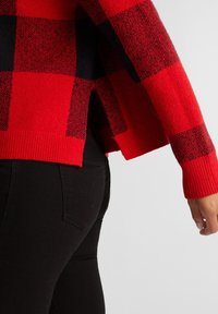 Suéter a cuadros rojo y negro con un corte holgado, puños acanalados y aberturas laterales. Textura suave visible, combinado con jeans ajustados negros.