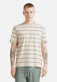 Camiseta de manga corta color beige con rayas grises, cuello redondo y un bolsillo en el pecho. El material parece suave, con un diseño de corte holgado.