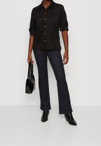 Svart mocka skjorta med knappar och två bröstfickor, kombinerad med mörka utsvängda jeans och svarta ankelstövlar, hållande en glänsande svart handväska.