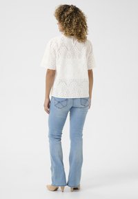Blusa bianca ricamata con maniche corte e a palloncino, decorata con motivi ad occhio di bue, abbinata a jeans flare azzurri e tacchi beige.