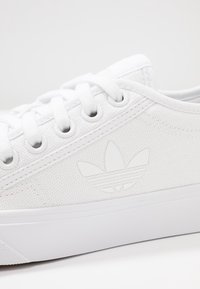 Vita tygskor med en texturerad yta, utrustade med runda vita öljetter, vita skosnören och en upphöjd vit logotyp på sidan.