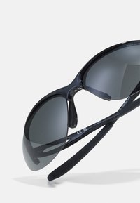 Juodi sportiniai saulės akiniai su pilkais objektyvais, išlenktu rėmo dizainu ir lanksčiomis kojelėmis. Išsiskiria elegantišku, blizgiu apdailos paviršiumi.