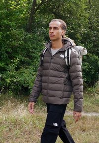 Rudas pūkuotas švarkas su horizontaliomis siūlėmis, pilkas marškinėlis, juodos krovinių kelnės ir kuprinė. Natūrali žalia fono aplinka su žole.