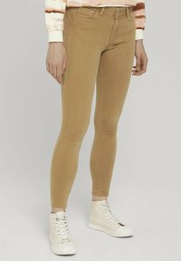 Persona vestida con jeans ajustados color beige y zapatillas altas blancas, de pie contra un fondo claro y liso.