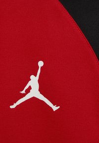 Röd tyg med en vit silhuett av en hoppande figur, med ett svart accentområde. Ytan är slät och sportig.