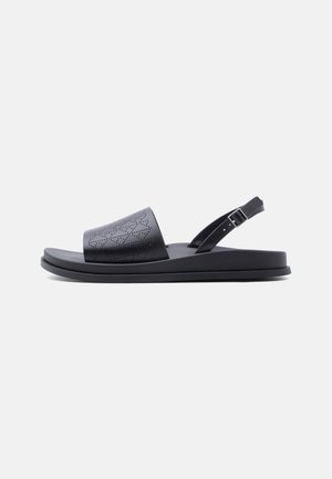 Black flat sandal with perforated wide front strap and adjustable buckle ankle strap on a plain light background.