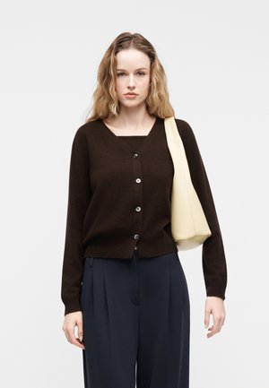 Žena s dlhými vlnitými vlasmi má na sebe tmavohnedý zapnutý kardigán, námornícke modré plisované nohavice a drží krémovú kabelku cez rameno na jednoduchom svetlom pozadí.