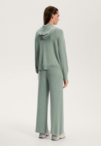 Hedvábně zelená ráčna pletená mikina a široké kalhoty z jemného, strukturovaného materiálu. Obsahuje kapuci a volný střih.