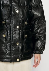Czarna puchowa kurtka z błyszczącym wykończeniem, z złotymi zatrzaskami, elastycznymi mankietami i przednimi kieszeniami. Tkanina o fakturze z pikowanym wzorem.