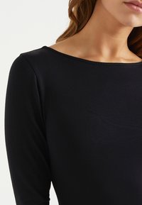 Kvinna iklädd en enkel svart långärmad topp med båturringning, som visar hennes nacke, axel och del av hennes läppar.