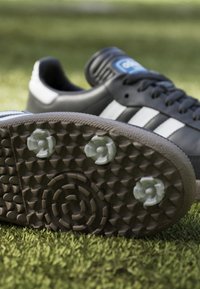 Czarny but sportowy z białymi paskami, teksturalna gumowa podeszwa z okrągłymi wzorami i sześcioma wyraźnymi wcięciami. Niski profil, solidna konstrukcja.