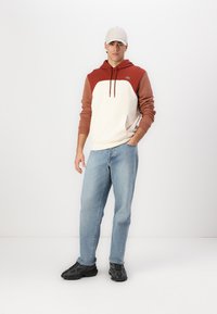 Felpa con cappuccio bianca e ruggine con tasca a marsupio, indossata con jeans azzurri chiari e sneakers nere. Il modello indossa un cappellino chiaro.