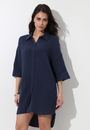 Donna con capelli scuri e mossi che indossa un abito navy blu con bottoni, maniche a tre quarti e taglio morbido, in piedi davanti a uno sfondo chiaro e uniforme.