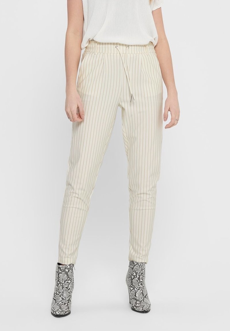 Femme portant un pantalon crème à fines rayures, coupe fuselée avec une taille à cordon, et des bottines à motif peau de serpent, debout sur fond blanc.