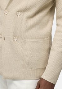 Beige dvouřadý blejzr vyrobený z texturované tkaniny, který má dva knoflíky a boční kapsu, s viditelnými švy na okrajích.