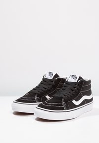 Schwarze High-Top-Sneaker aus Wildleder mit weißen Akzenten, versehen mit einem kontrastierenden Seitenstreifen, runden Schnürsenkeln und einem gepolsterten Kragen.