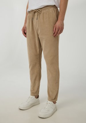 Pantaloni in corda beige con una vestibilità rilassata, dotati di una vita elasticizzata e chiusura con lacci. Indossati con sneakers bianche.