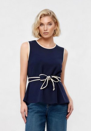 Blonde Frau trägt ärmelloses marineblaues Oberteil mit weißem Saum und gebundenem Gürtel, kombiniert mit blauer Jeans, steht vor einem einfarbigen Hintergrund.