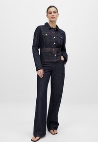 Temno modra jakna iz denima s kontrastnimi rdečimi šivi, gumbnimi zapiralci in dvema žepoma na prsih; v kombinaciji z wide-leg hlačami iz ustreznega materiala.