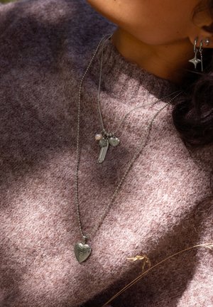 Colliers superposés en argent sur un pull en laine rose doux et texturé ; avec un pendentif en forme de cœur, un charme en forme de clé et une accent en perle.