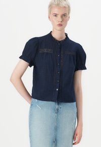 Mørkeblå kortermet bluse med volangkrage, blonder og frontlukking med knapper, kombinert med en lys blå denimskjørt.