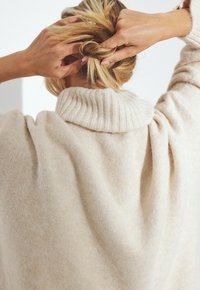 Beige oversized sweater with a ribbed turtleneck, soft texture, and loose fit, showing rolled sleeves and a high neckline.